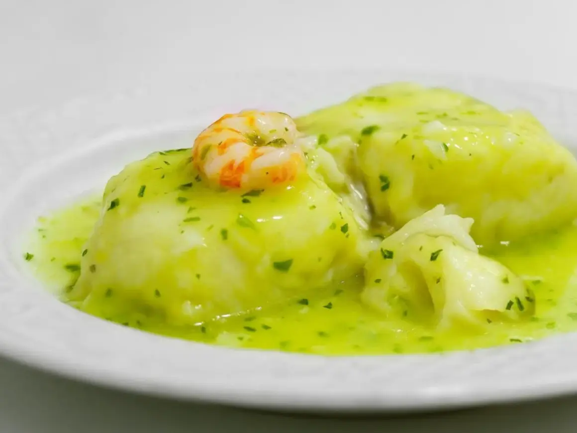 Merluza en salsa verde, un plato tradicional con trozos de pescado blanco y un camarón encima, bañado en una deliciosa salsa verde.