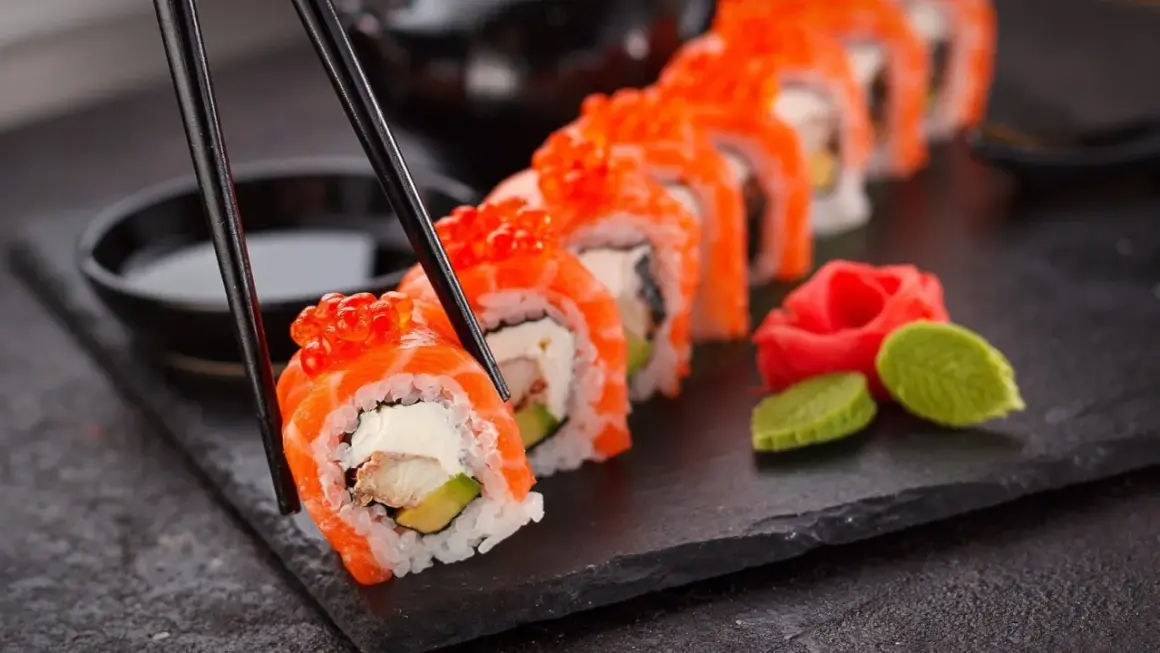 Palillos recogen un rollo de sushi con salmón y huevas de salmón, listo para disfrutar. Aprende como hacer sushi.