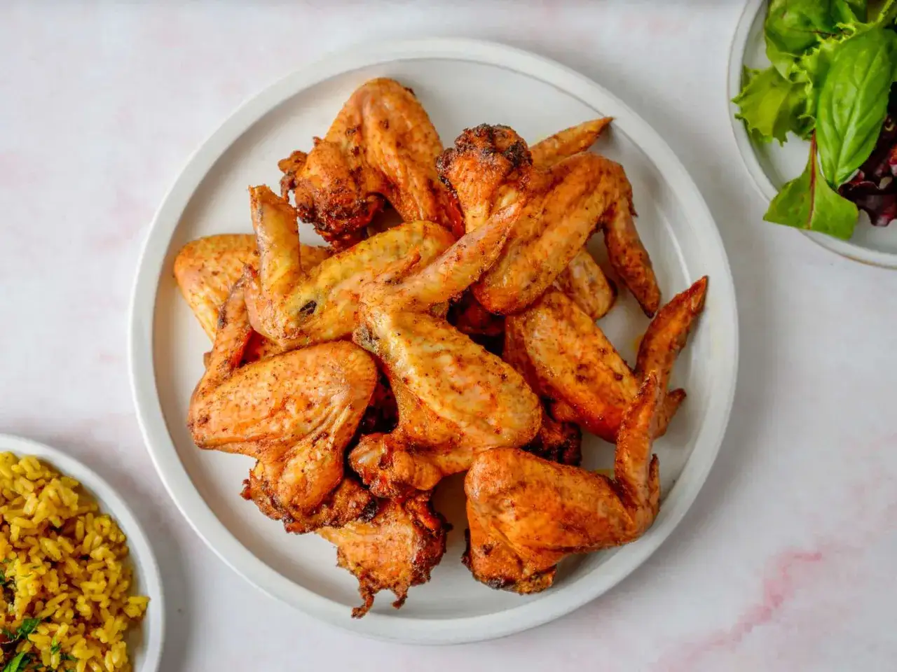 Un plato blanco lleno de alitas de pollo al horno, doradas y crujientes, listas para disfrutar. Acompa&ntilde;adas de arroz amarillo y una ensalada fresca.