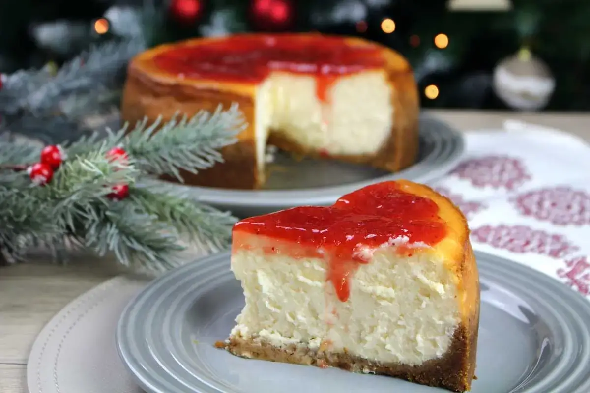 Un delicioso new york cheesecake con cobertura de fresa, servido en un plato gris, con un árbol de Navidad de fondo.