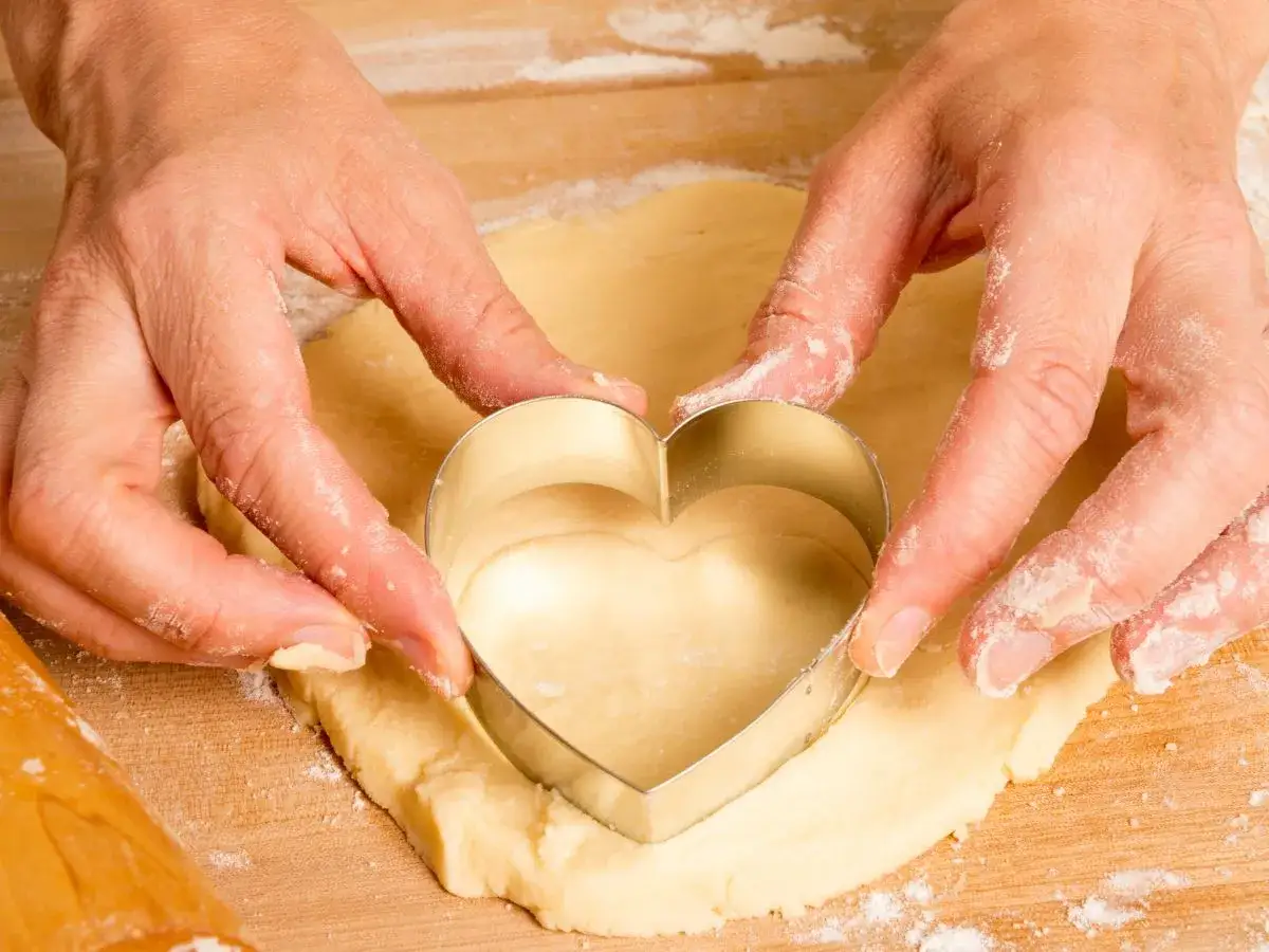 Manos cortan masa de galletas con un cortador en forma de coraz&oacute;n.