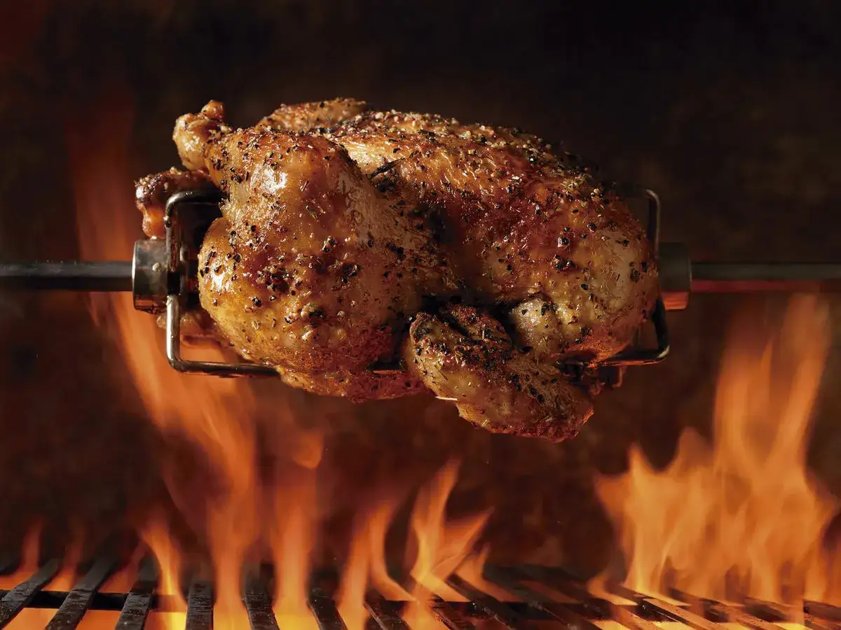 Pollo al ast dorado y jugoso girando sobre brasas ardientes, con llamas anaranjadas envolviendo la parrilla.