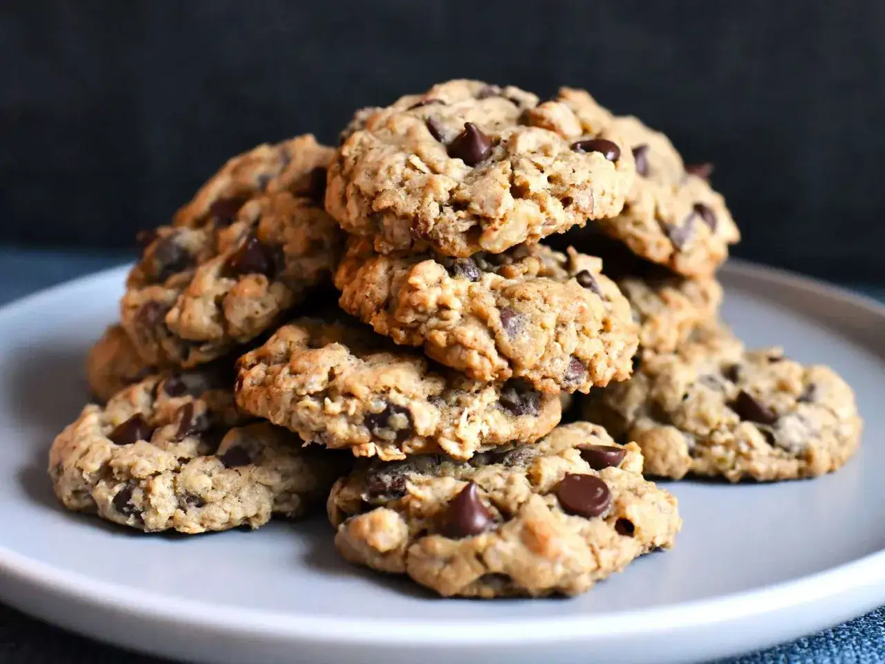 Mont&oacute;n de galletas de avena y chocolate reci&eacute;n horneadas, con trozos de chocolate derretido, apiladas en un plato gris.