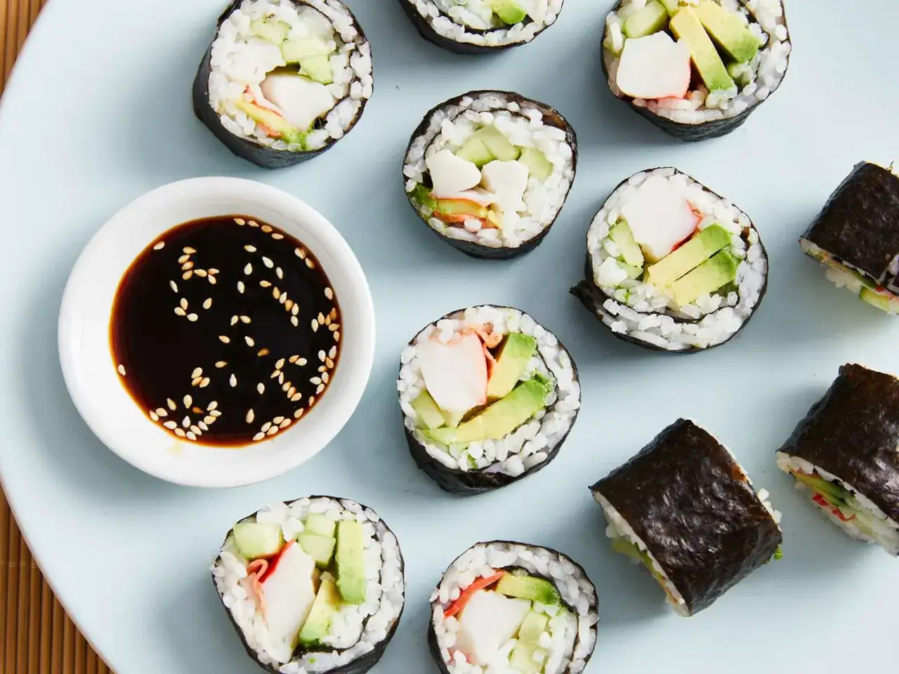 Rollos de sushi caseros con aguacate, pepino y surimi, listos para aprender como hacer sushi. Salsa de soja con s&eacute;samo.