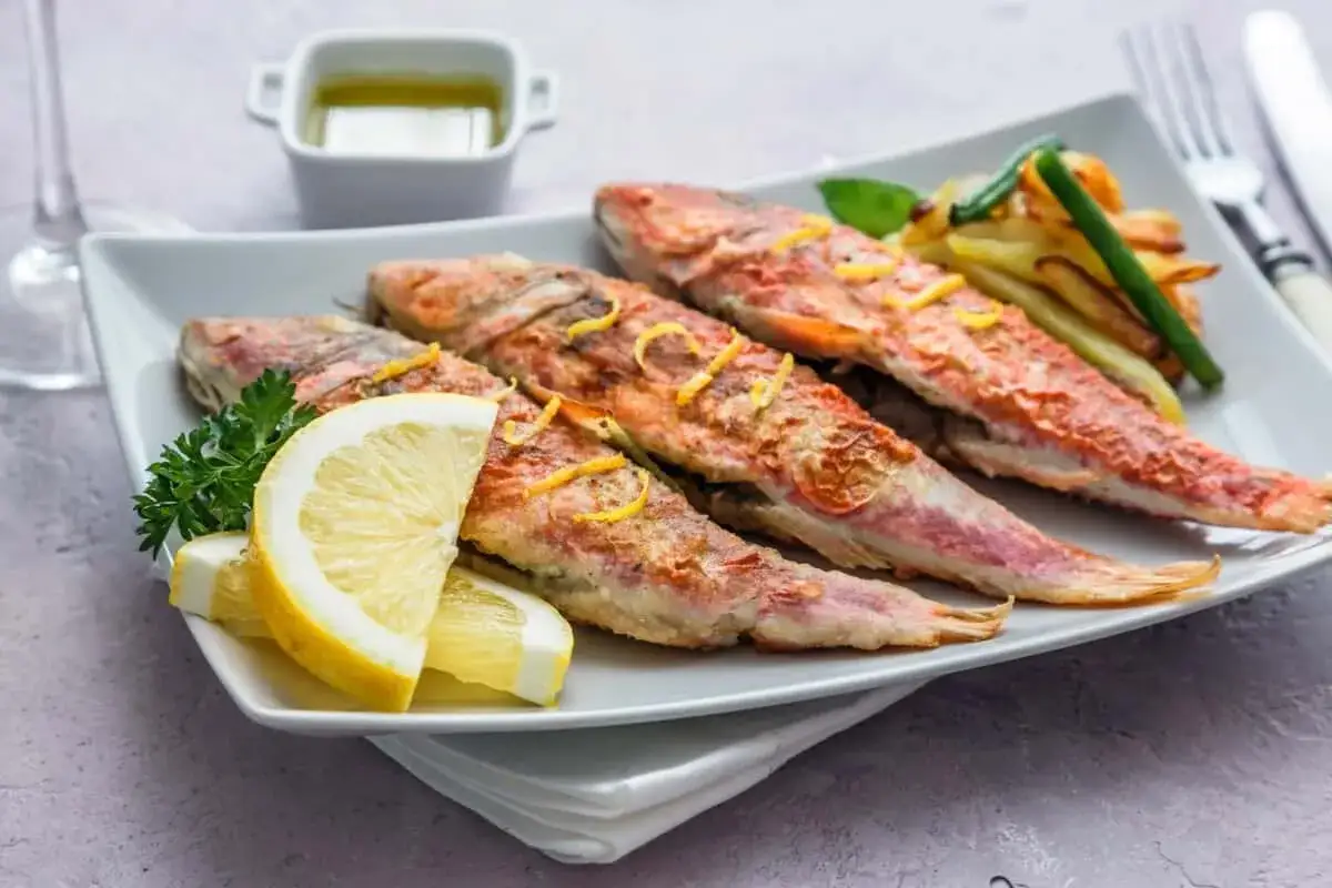 Cuatro salmonetes al horno, dorados y jugosos, servidos con rodajas de limón, perejil y patatas fritas.