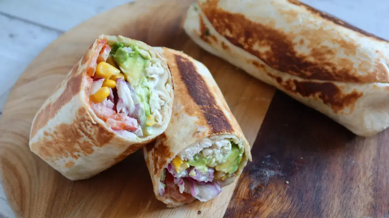 Burritos de pollo reci&eacute;n hechos, rellenos de aguacate, ma&iacute;z, tomate y cebolla morada, servidos en una tabla de madera.