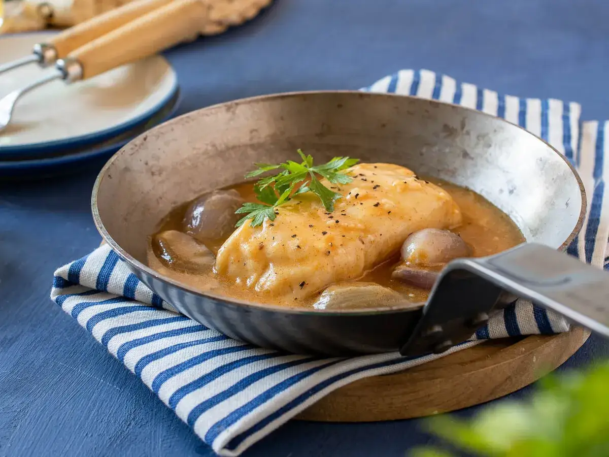 Merluza en salsa con cebollitas, una de las mejores recetas merluza congelada. Servida en sartén metálica sobre mantel a rayas.