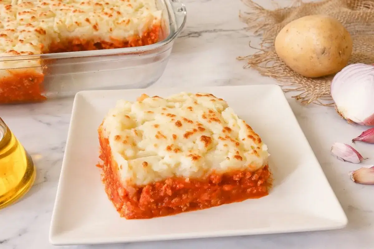 Porci&oacute;n de pastel de carne casero, con pur&eacute; de patatas dorado y relleno jugoso. Ingredientes frescos como patatas y cebolla en el fondo.