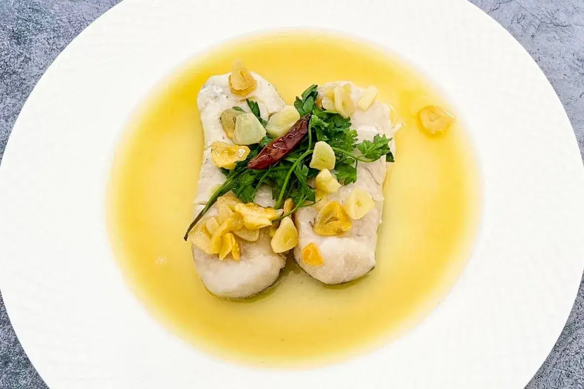 Dos lomos de merluza al pil pil, con l&aacute;minas de ajo dorado, perejil fresco y un toque de guindilla, servidos en un plato blanco.