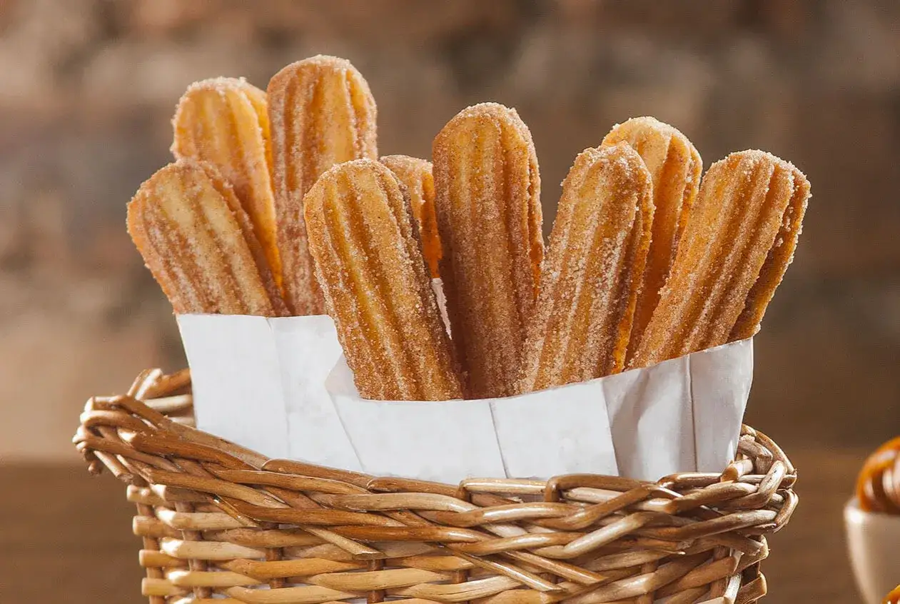 Cesta de mimbre llena de churros sin gluten espolvoreados con az&uacute;car y canela. &iexcl;Un placer crujiente para todos!