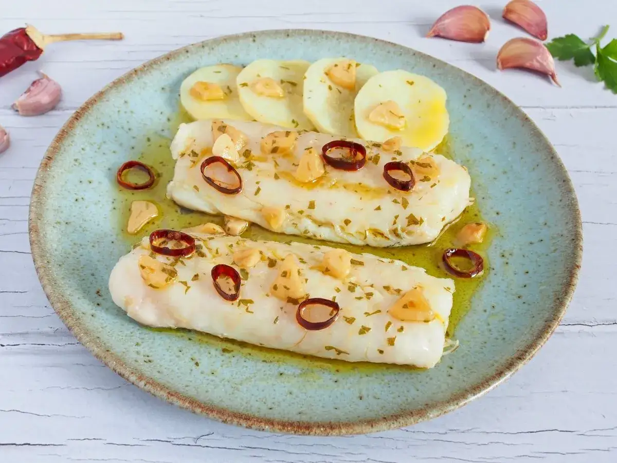 Dos lomos de merluza a la bilbaina, con patatas, ajo laminado y guindilla, servidos en plato azul.