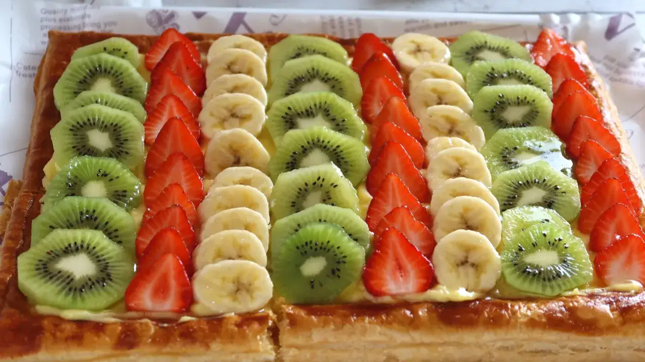 Tarta de frutas con filas de fresas, rodajas de pl&aacute;tano y kiwi sobre una base dorada y cremosa.
