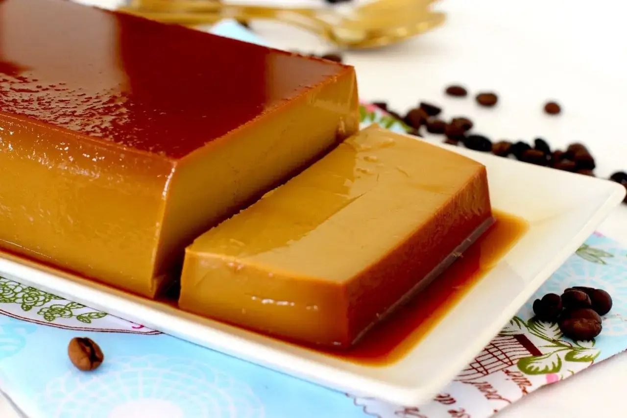 Un delicioso flan de caf&eacute;, con su caramelo brillante, servido en una bandeja blanca, rodeado de granos de caf&eacute; tostado.