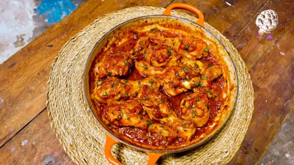 Sabroso pollo al chilindrón en cazuela naranja, servido sobre una esterilla de mimbre en una mesa de madera rústica.