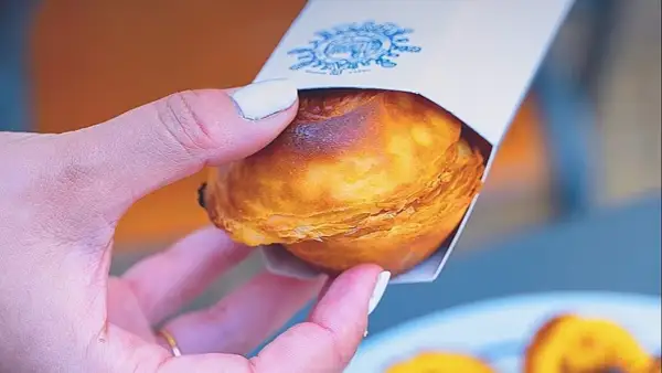 Mano sosteniendo un pastelito de Belém recién horneado, con su masa hojaldrada dorada y un toque de caramelo.