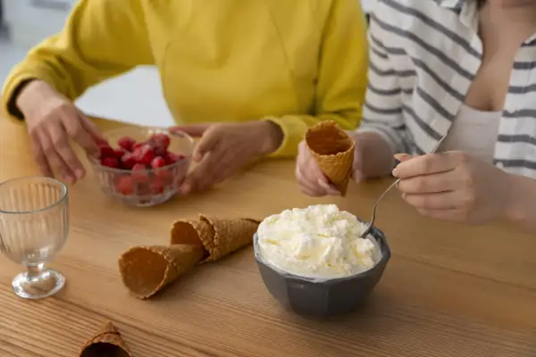 Dos personas preparan helado casero. Hay un tazón de helado cremoso, fresas, conos de helado y un vaso.