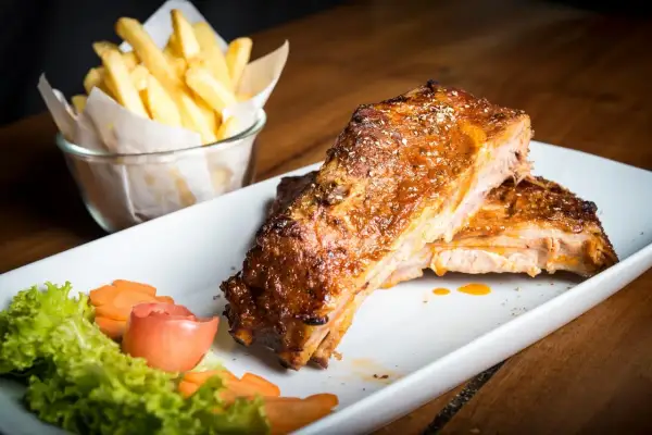 Costillas al horno jugosas y doradas, servidas con papas fritas crujientes y una guarnición fresca de lechuga, zanahoria y tomate.