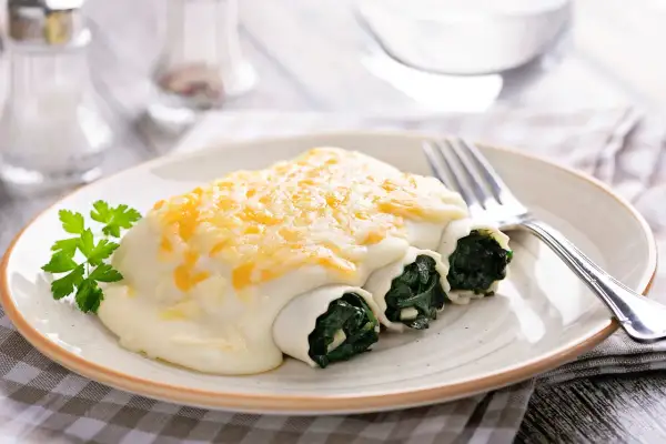 Canelones de espinacas cubiertos con bechamel y queso gratinado, servidos en un plato con perejil fresco.