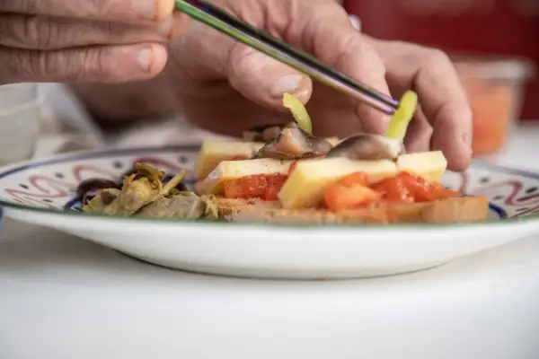 Manos preparan pa amb oli con queso, tomate, aceitunas y alcachofas en una tostada.
