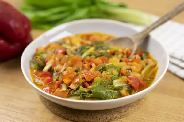 Un plato humeante de guiso de verduras, ideal para recetas con acelgas. Contiene trozos de tomate, pimiento rojo y hojas verdes.