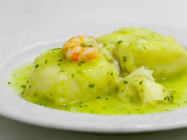 Merluza en salsa verde, un plato tradicional con trozos de pescado blanco y un camarón encima, bañado en una deliciosa salsa verde.